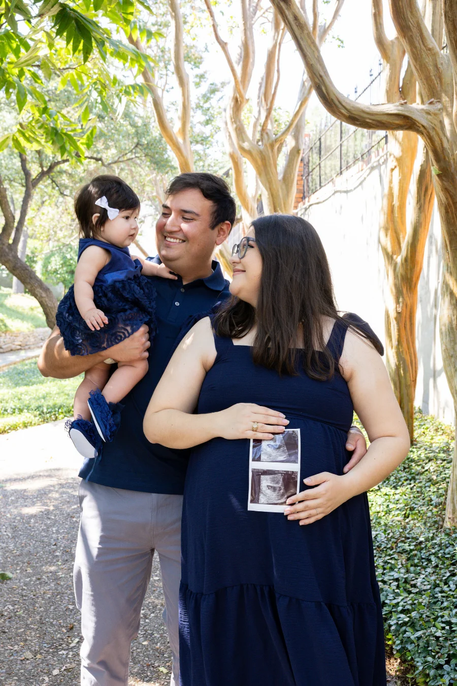 A pregnant mother holds an ultrasound print to her belly as the father, holding their baby daughter, smiles beside her