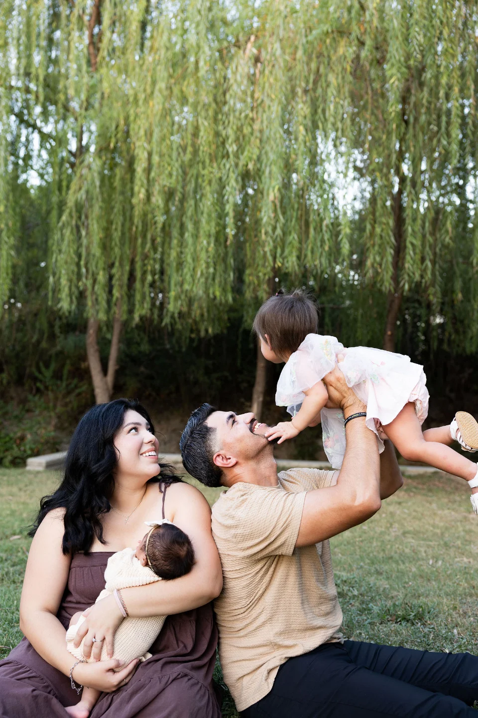 Under a willow tree, a father lifts his laughing toddler overhead while the mother sits beside him holding their newborn