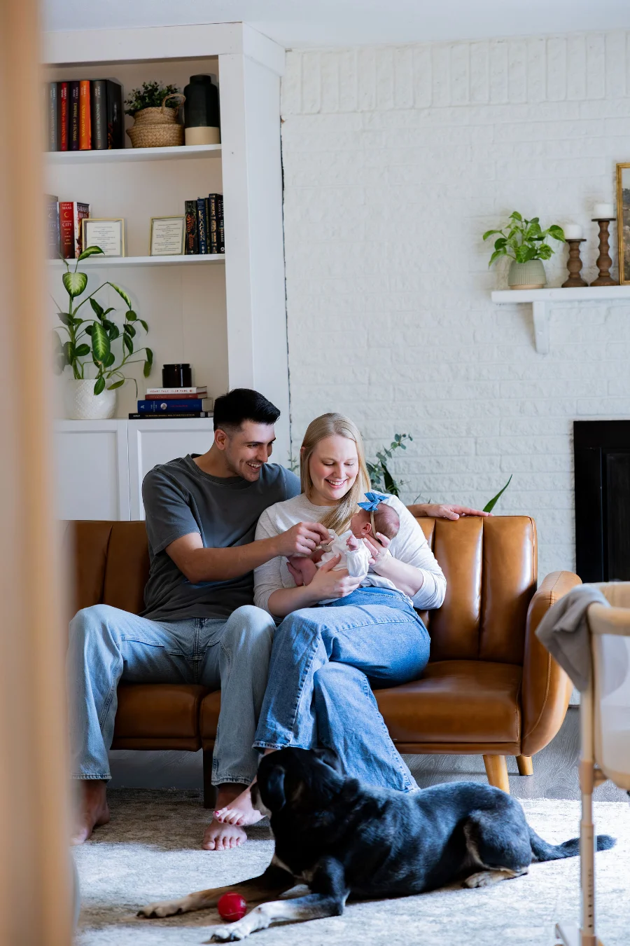 New parents smile down at their newborn daughter on a leather sofa at home while their dog lies on the rug in front of them