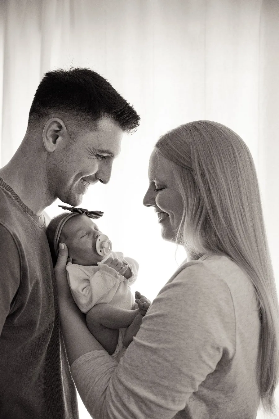 Backlit by a bright window, new parents smile down at their sleeping newborn daughter, in black and white