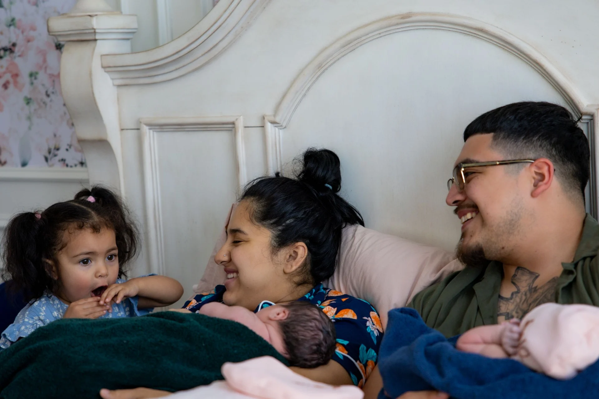 The Meza mother laughs in bed with one of her newborn twins on her chest, the father holding the other twin and their toddler looking on in wonder