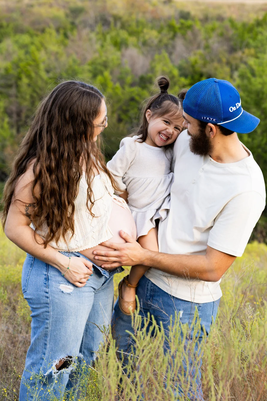 A father rests his hand on his pregnant partner's bare belly as their laughing daughter hugs him in a grassy field
