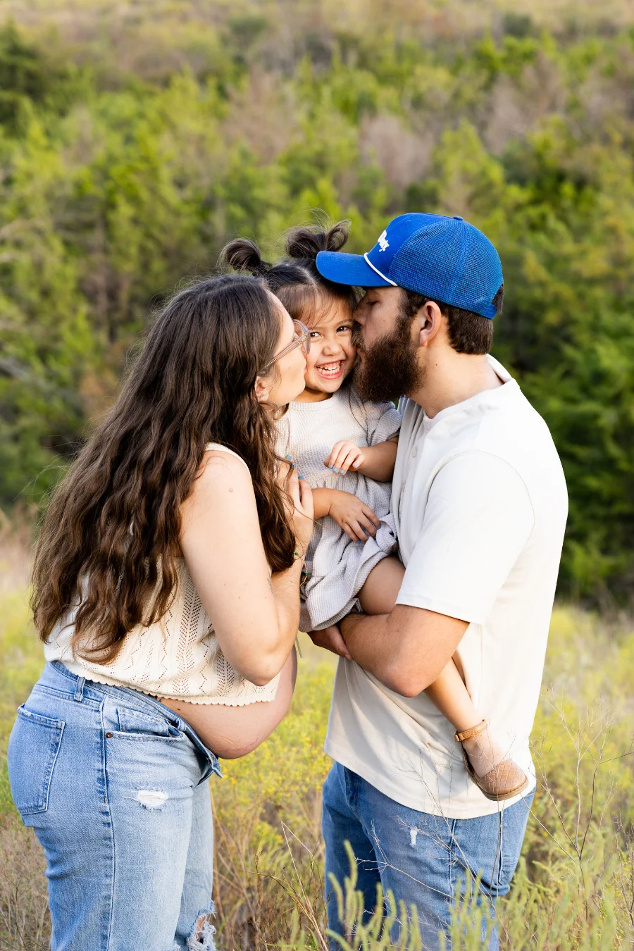 A pregnant mother and her husband kiss their laughing daughter's cheeks in a grassy field