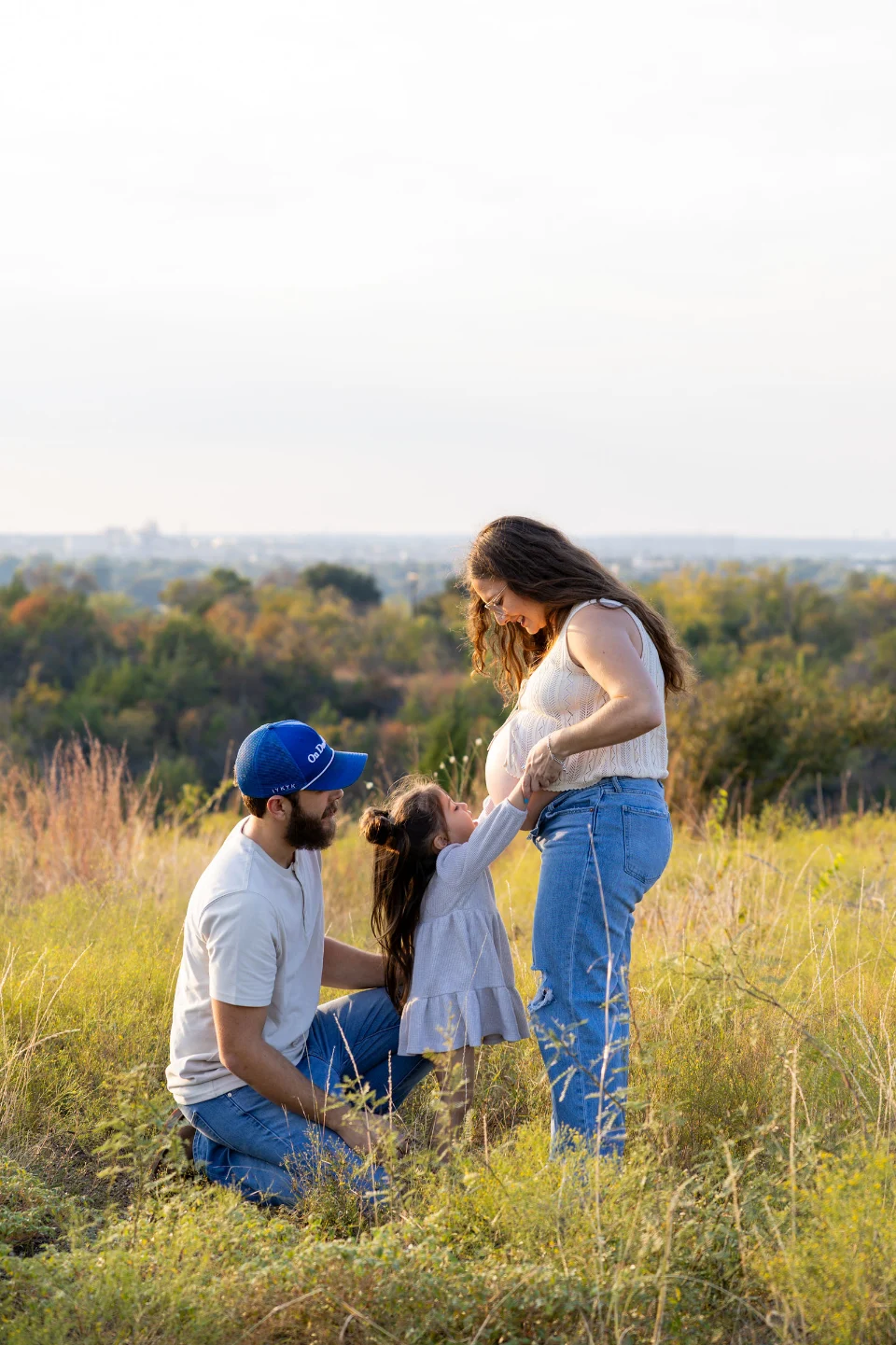 A little girl holds her pregnant mother's hands and looks up at her belly while her father kneels beside them in tall grass