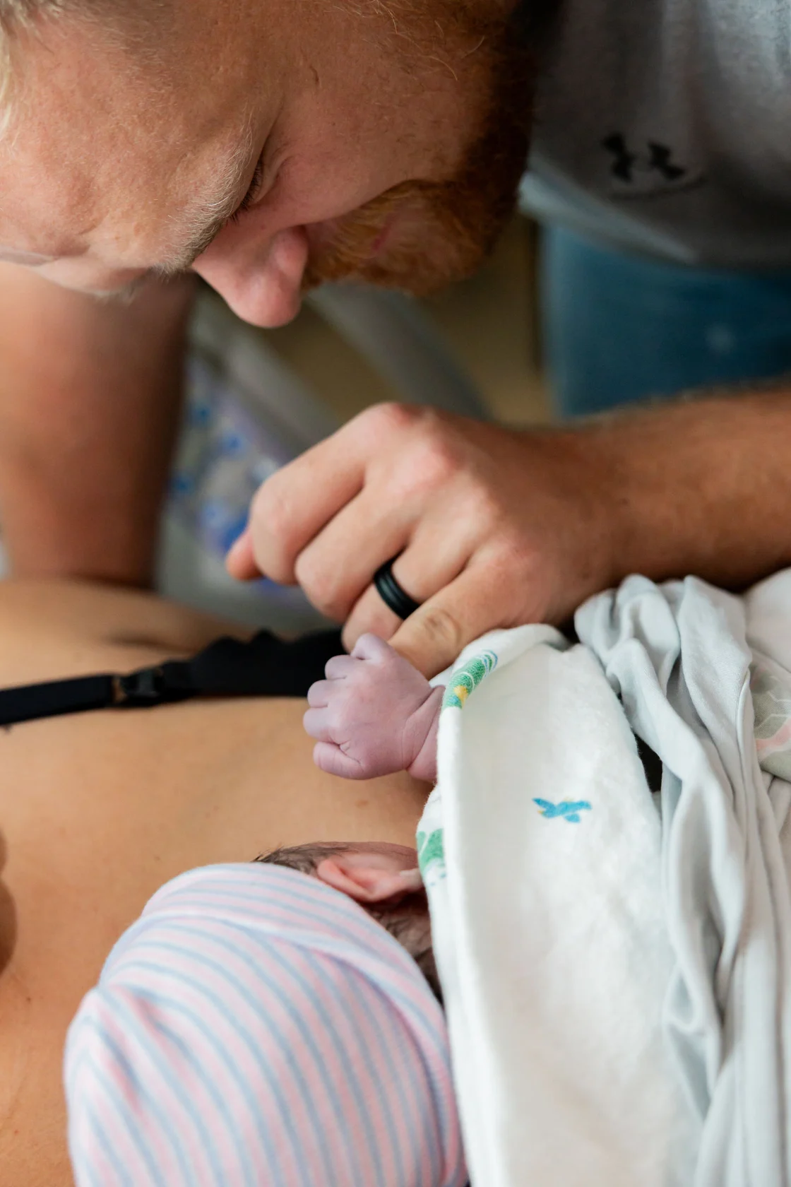Minutes after birth in the hospital, a father's hand holds his newborn's tiny fist as the baby rests on the mother's chest
