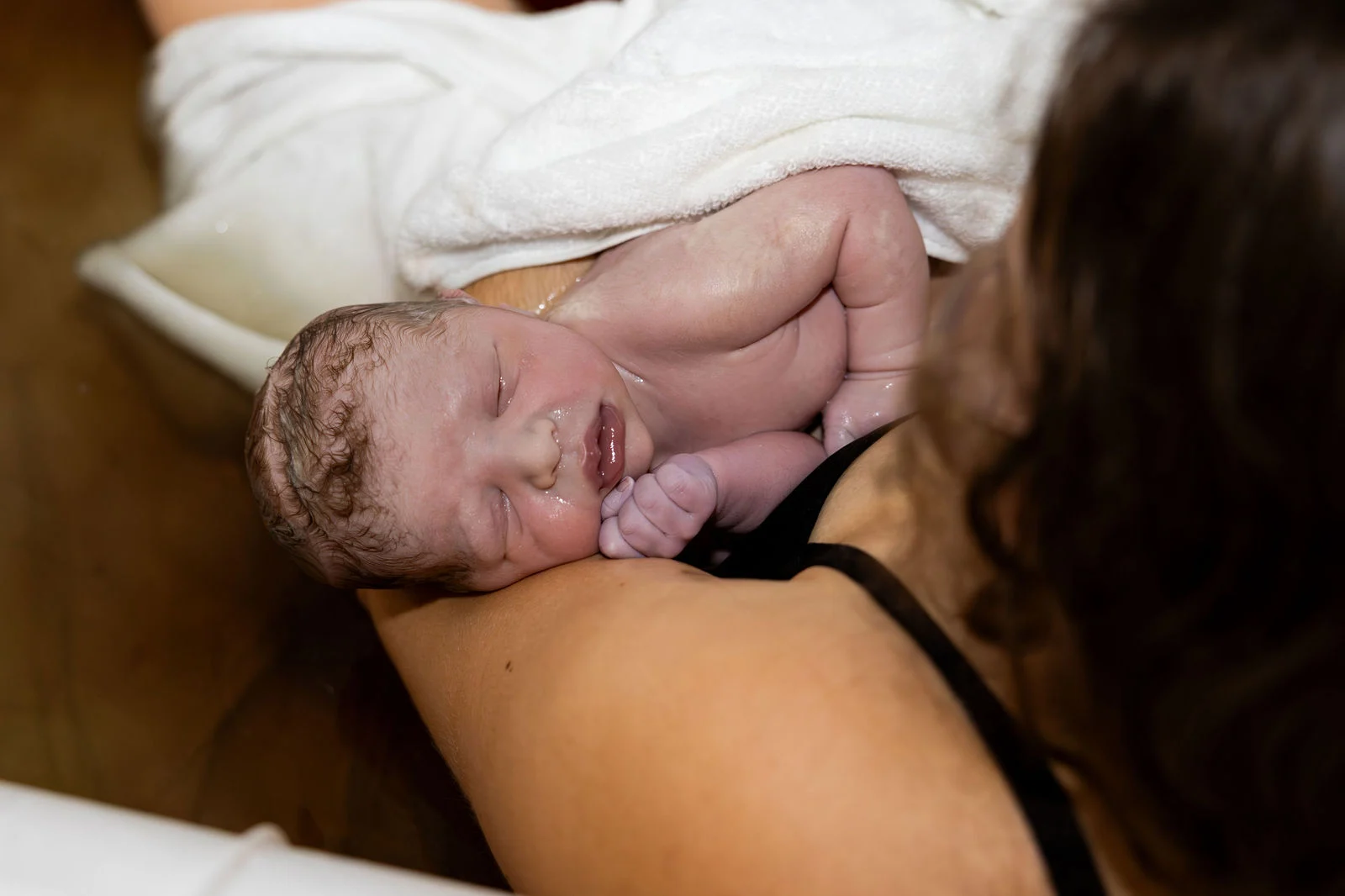 Seconds after a water birth at home, a newborn rests skin to skin on the mother's shoulder, one tiny fist tucked under the chin