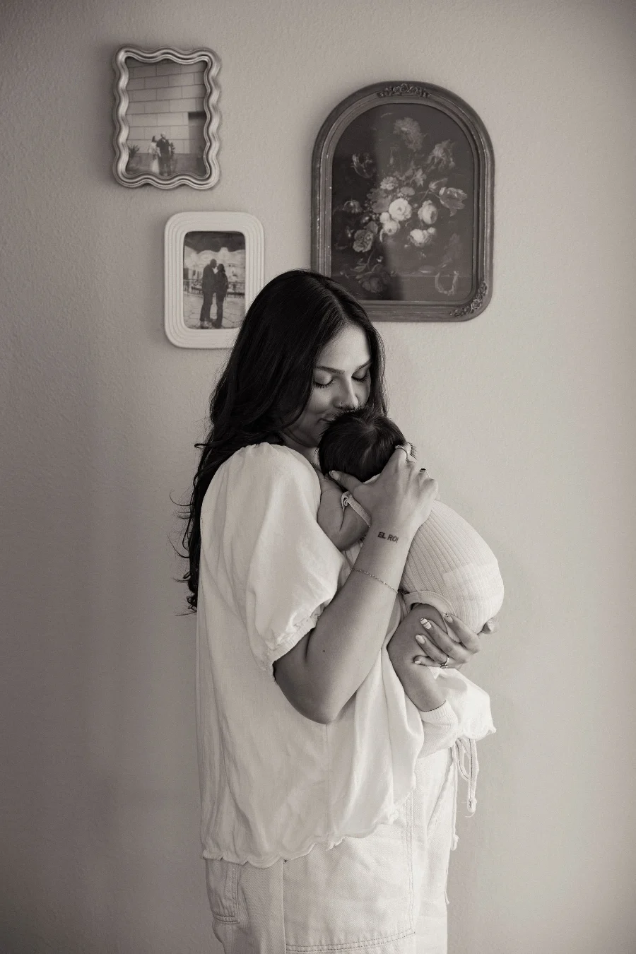 A mother with her eyes closed kisses the head of the newborn she holds to her chest, beside a wall of framed pictures, in black and white