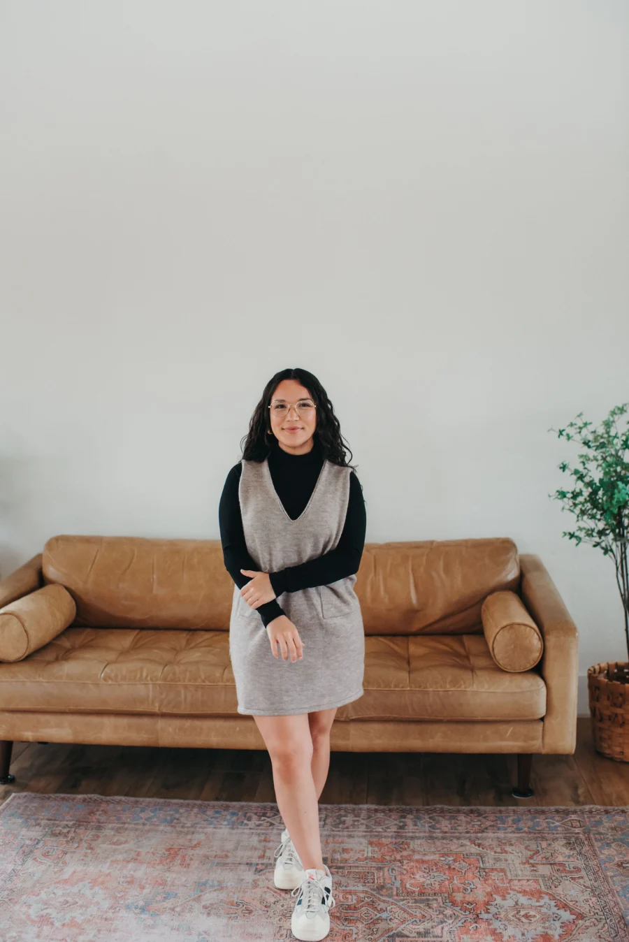 Jordan Flores in glasses and a gray sweater dress, standing in front of a leather sofa