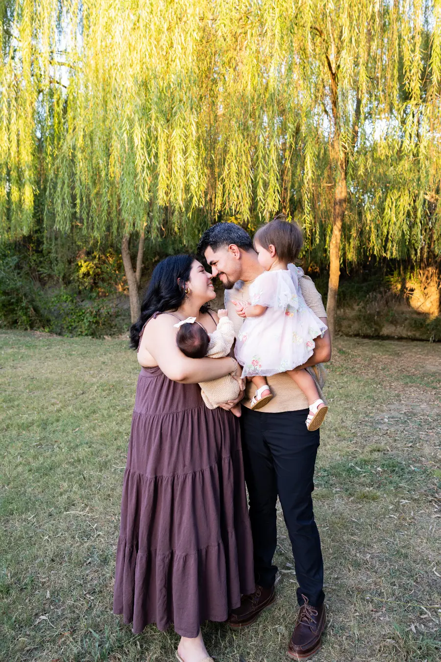 Under a willow tree, parents touch noses while the mother holds their newborn and the father holds their toddler daughter, both little ones facing away